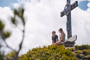 Zwei Frauen sitzen lächelnd auf einem Berg neben dem Gipfelkreuz. 