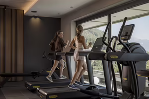 Zwei Frauen joggen auf Laufbändern in einem modernen Fitnessraum mit großem Fenster und Blick auf die Berge.