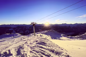 Eine Berglandschaft mit Schnee, einem Skilift und der aufgehenden Sonne über den Bergen.