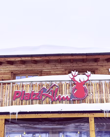 Platzlalm Schriftzug bei Schneefall Die Platzlalm mit einem großen, rot-weißen Schild "Platz Alm" und einem Hirsch-Logo. Schnee auf dem Dach.