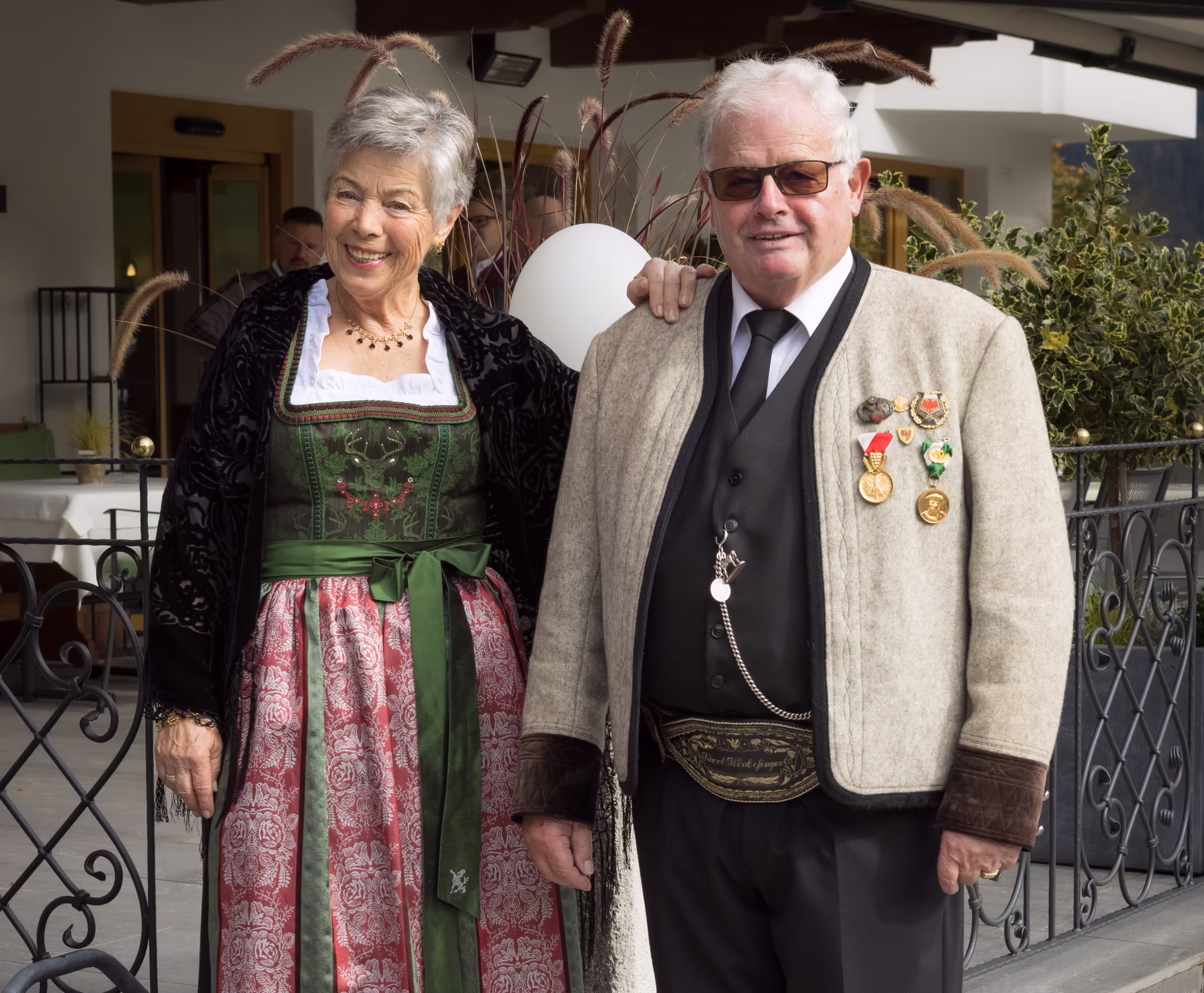 Anni und Karl Wimpissinger – Seniorchefs im Platzlhof Seniorchefs in traditioneller Kleidung stehen zusammen vor dem Hotel Platzlhof. Oma Anni trägt ein Dirndl, Opa Karl trägt einen Tuxer.