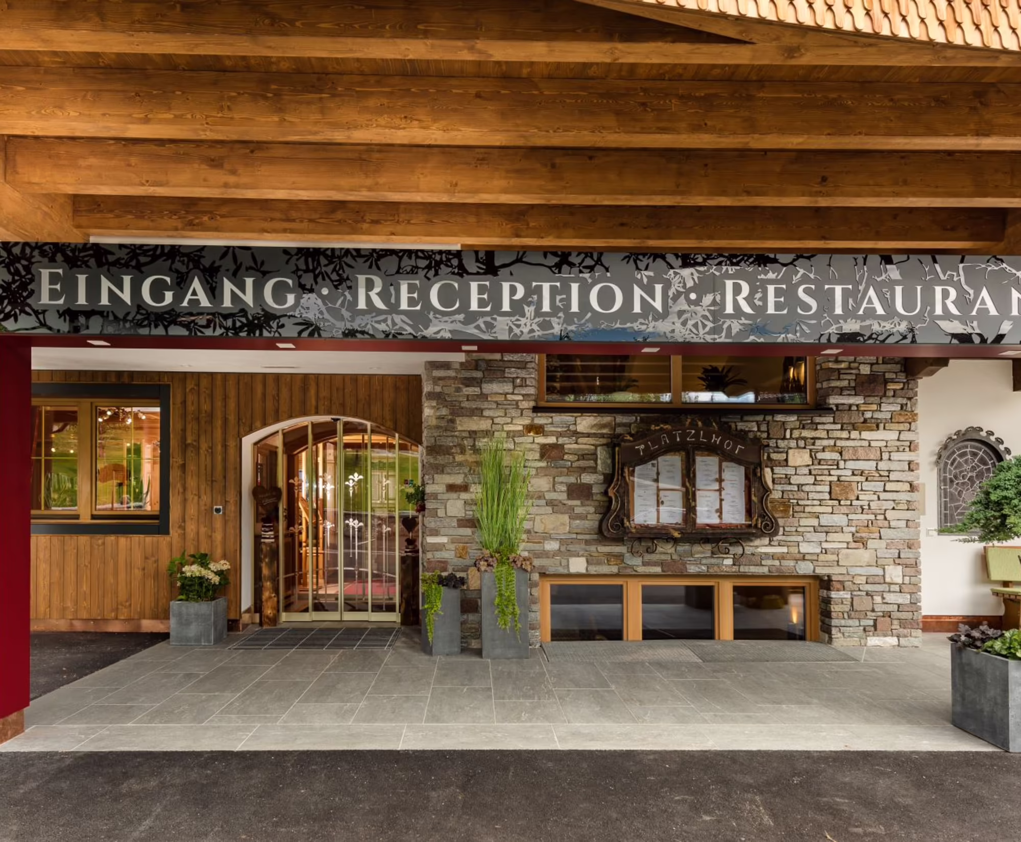 Eingang Rezeption Hotel Platzlhof Ried Eingang vom Hotel Platzlhof, das die Bereiche Rezeption und Restaurant umfasst. Eine Steinwand und Pflanzen in Töpfen sind sichtbar.