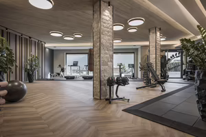 Moderne Fitnessstudio mit verschiedenen Geräten, Holzfußboden, Pflanzen und großen Fenstern, die viel Licht einlassen.
