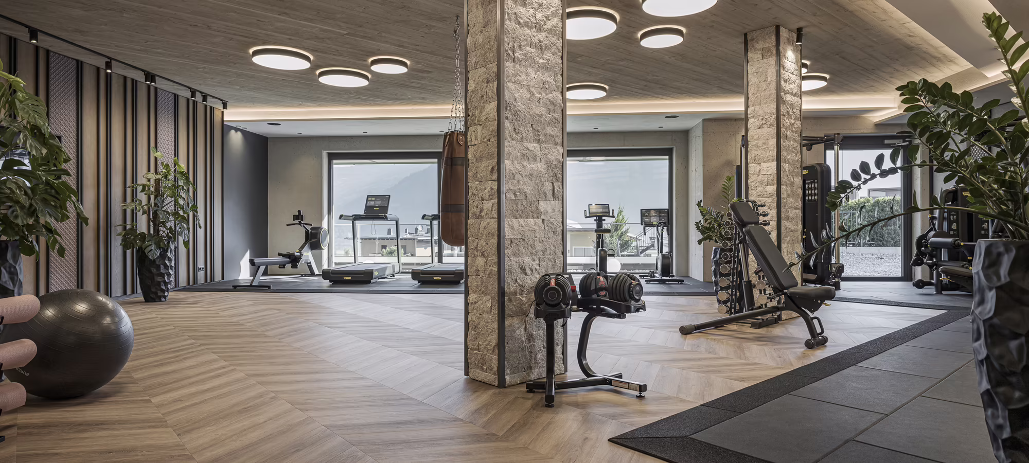 Moderne Fitnessstudio mit verschiedenen Geräten, Holzfußboden, Pflanzen und großen Fenstern, die viel Licht einlassen.