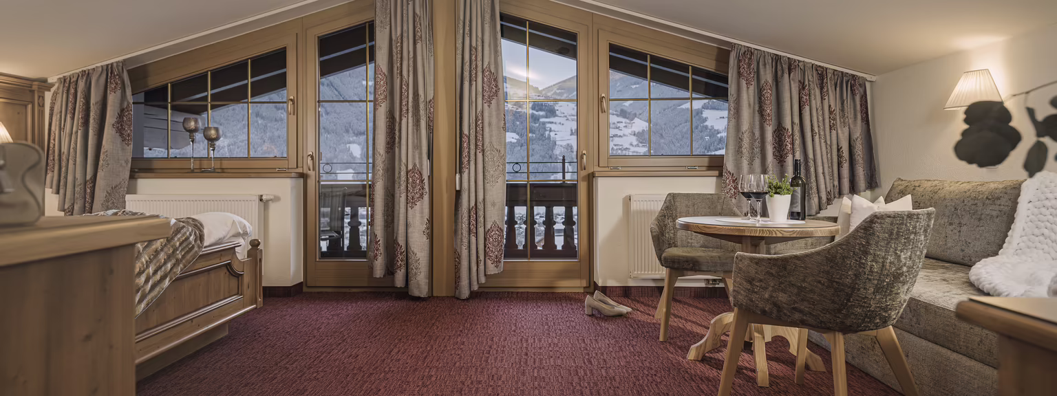 Gemütliches Zimmer mit Panoramafenstern, Teppichboden, einem Bett, einem Sofa und einem Tisch, umgeben von Bergen.