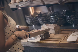 Eine Person schneidet Brot mit einem großen Messer auf einem Holzbrett in einer Bäckerei. Im Hintergrund sind weitere Brote sichtbar.