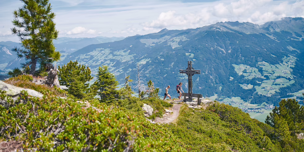 Wandern im Hochzillertal – Gipfelkreuz über dem Zillertal Wanderer stehen an einem Gipfelkreuz mit Aussicht auf grüne Täler und Berge unter einem klaren Himmel.
