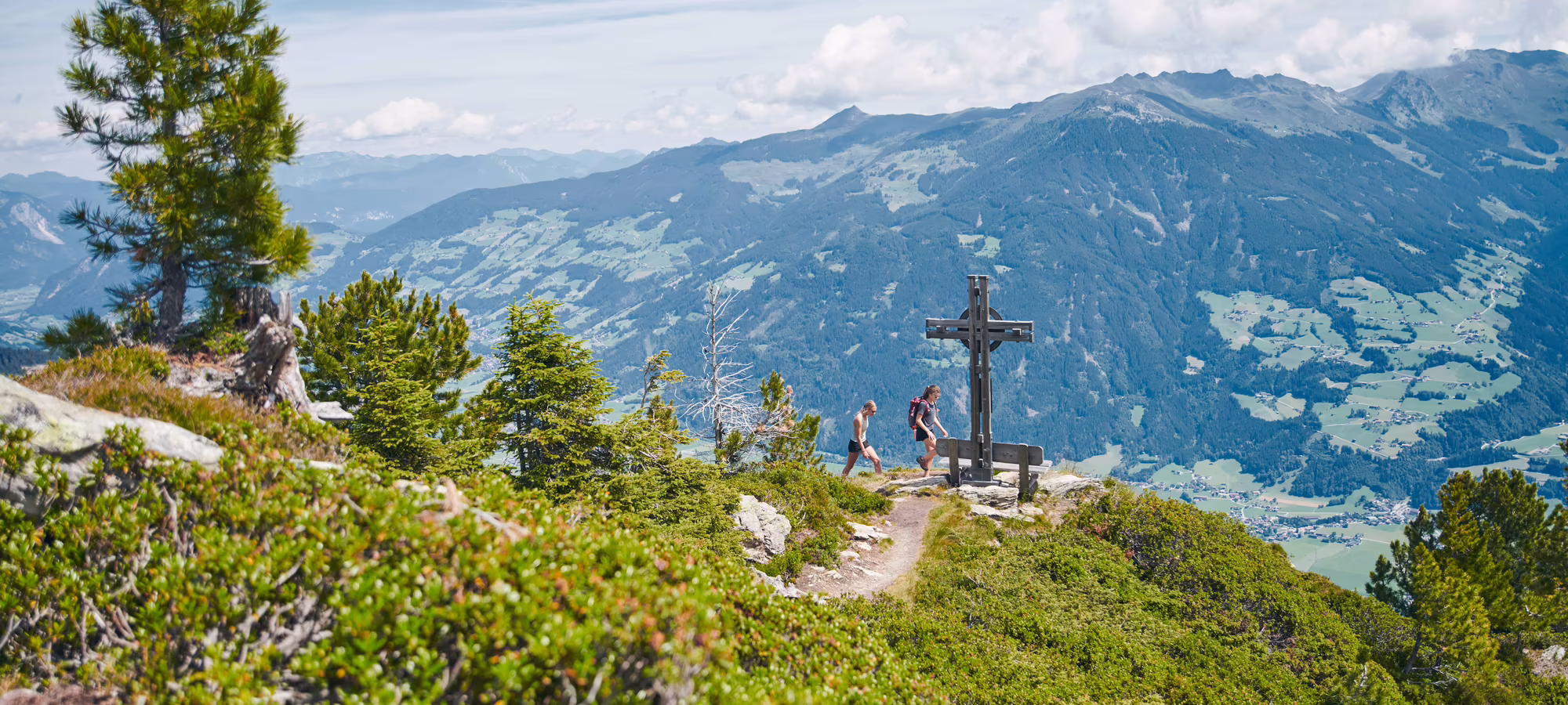 Wanderer stehen an einem Gipfelkreuz mit Aussicht auf grüne Täler und Berge unter einem klaren Himmel.