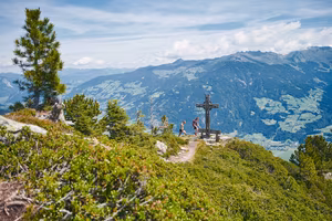 Wanderer stehen an einem Gipfelkreuz mit Aussicht auf grüne Täler und Berge unter einem klaren Himmel.