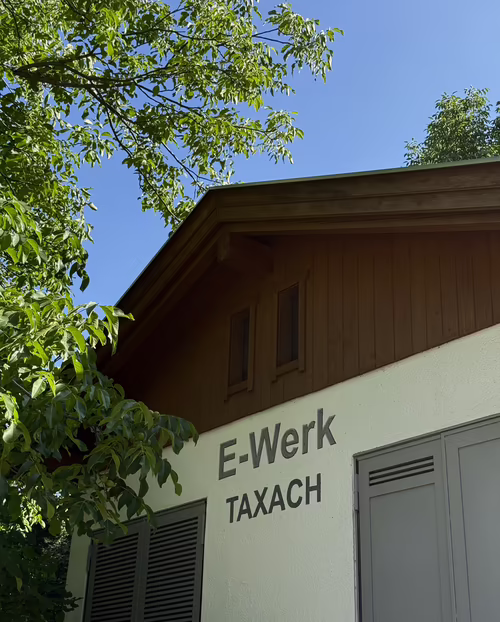 Das hauseigene Wasserkraftwerk der Familie Wimpissinger E-Werk Gebäude in Taxach, umgeben von Bäumen und blauem Himmel.
