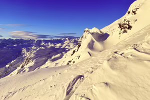 Schneebedeckte Berge unter einem klaren blauen Himmel, mit sanften Wellen und Felsen im Vordergrund.