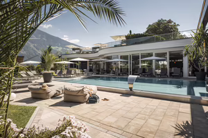 Ein moderner Pool-Bereich mit Liegen, Sonnenschirmen und umgeben von Pflanzen, vor einem Gebäudekomplex und Berglandschaft im Hintergrund.