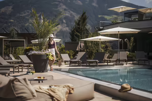 Eine Frau spaziert entspannt entlang des Außenpools im Hotel Platzlhof mit Sonnenliegen und Zillertaler Bergkulisse.