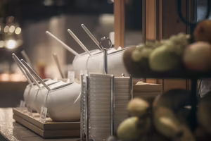 Weißes Geschirr mit Löffeln auf einem Buffet, daneben eine Obstschale mit bunten Früchten.