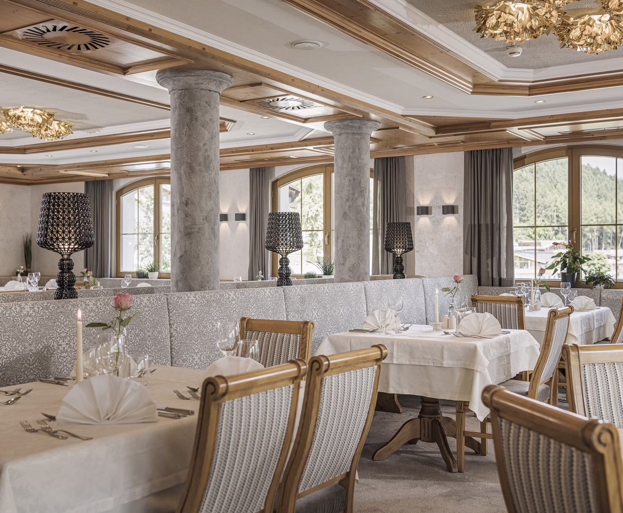Weiß gedeckte Tische mit Kerzen und Rosen im Restaurant des Hotel Platzlhof Elegantes Restaurant mit dekorierten Tischen, Stühlen und einem stilvollen Ambiente, beleuchtet von Kronleuchtern und Lampen.