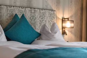 Ein Bett mit weißer Bettwäsche und türkisfarbenen Kissen vor einer dekorativen Kopfteilwand und einem Wandlicht.