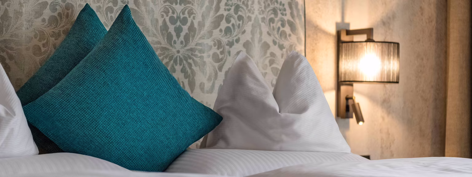 Entspannte Atmosphäre im Schlafzimmer Ein Bett mit weißer Bettwäsche und türkisfarbenen Kissen vor einer dekorativen Kopfteilwand und einem Wandlicht.