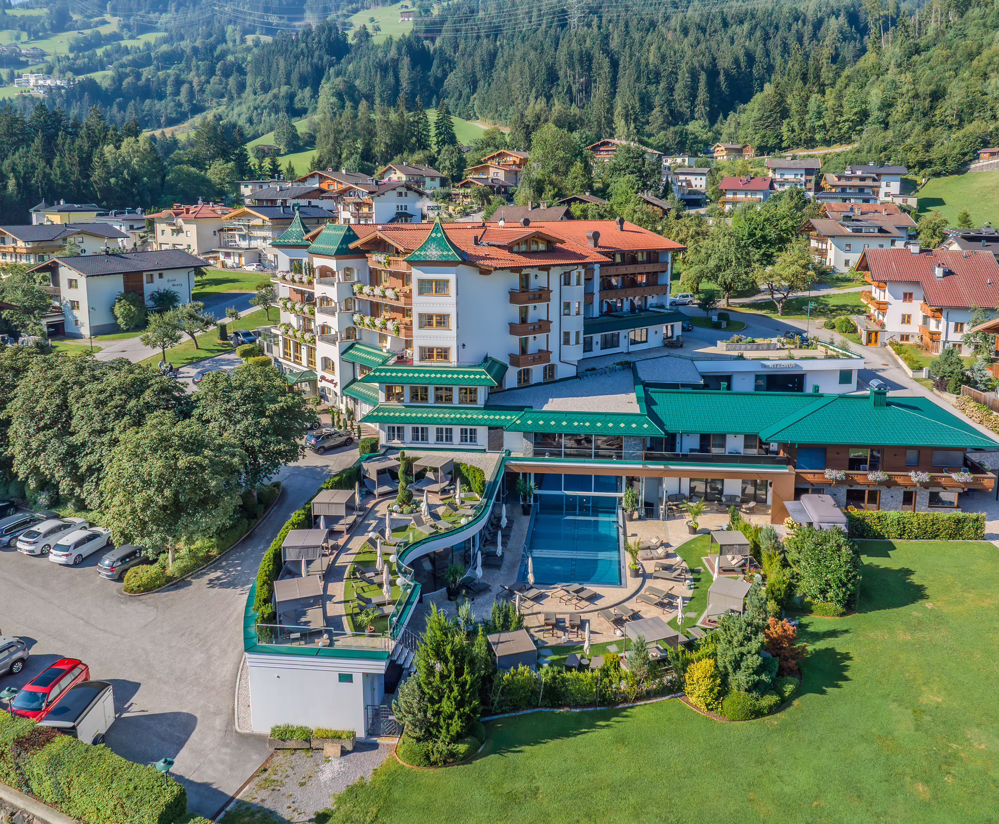 Drohnenaufnahme Hotel Platzlhof © level26 Luftaufnahme Hotel Platzlhof mit grünem Dach, umgeben von Bäumen und anderen Gebäuden in einer hügeligen Landschaft. Im Vordergrund sind Liegen und ein Poolbereich zu sehen.