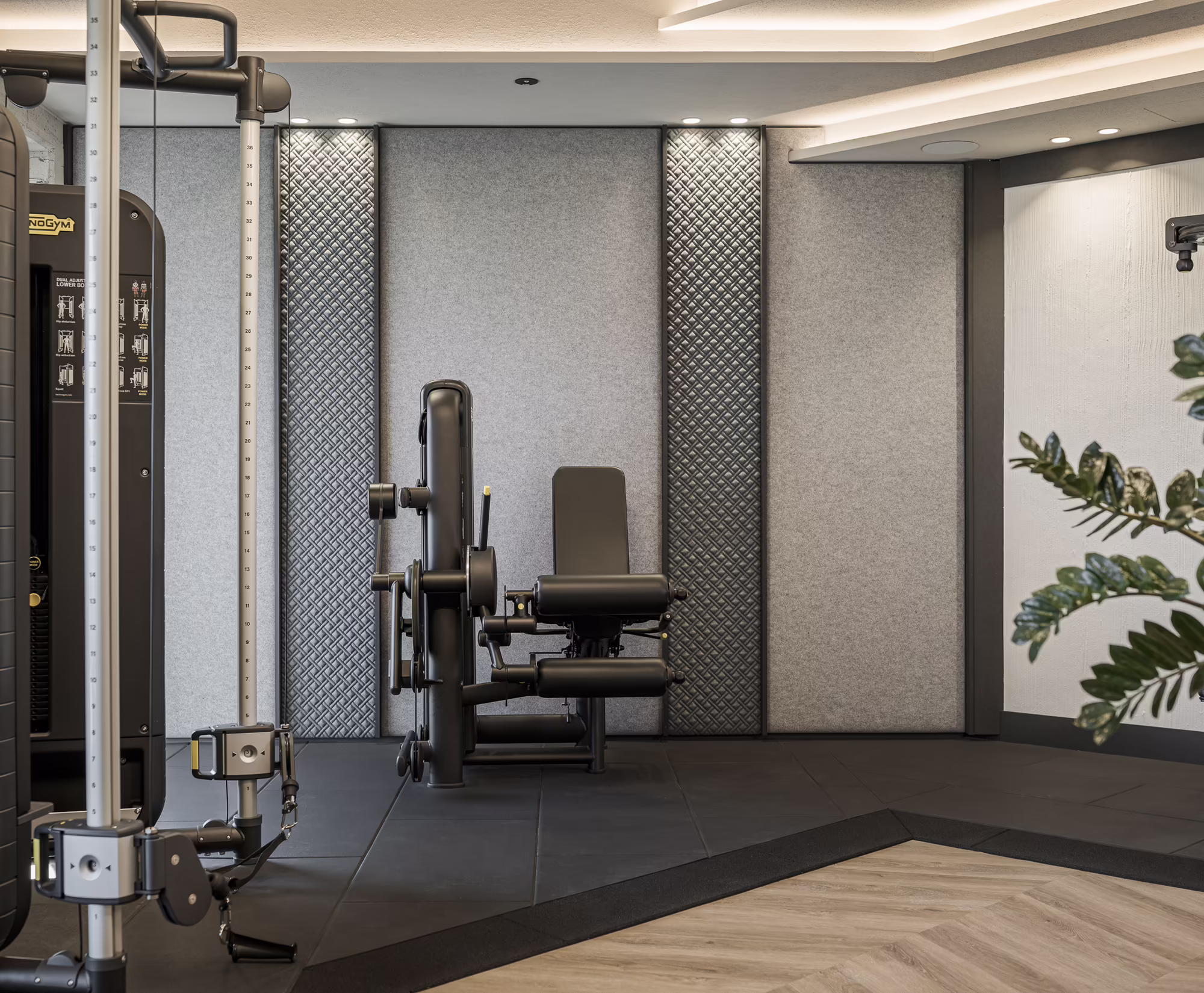 Krafttraining im Hotel Platzlhof Moderner Fitnessraum mit Trainingsgeräten, einer Hantelbank und dekorativer Wandgestaltung. Grünes Blattwerk ergänzt die Atmosphäre.