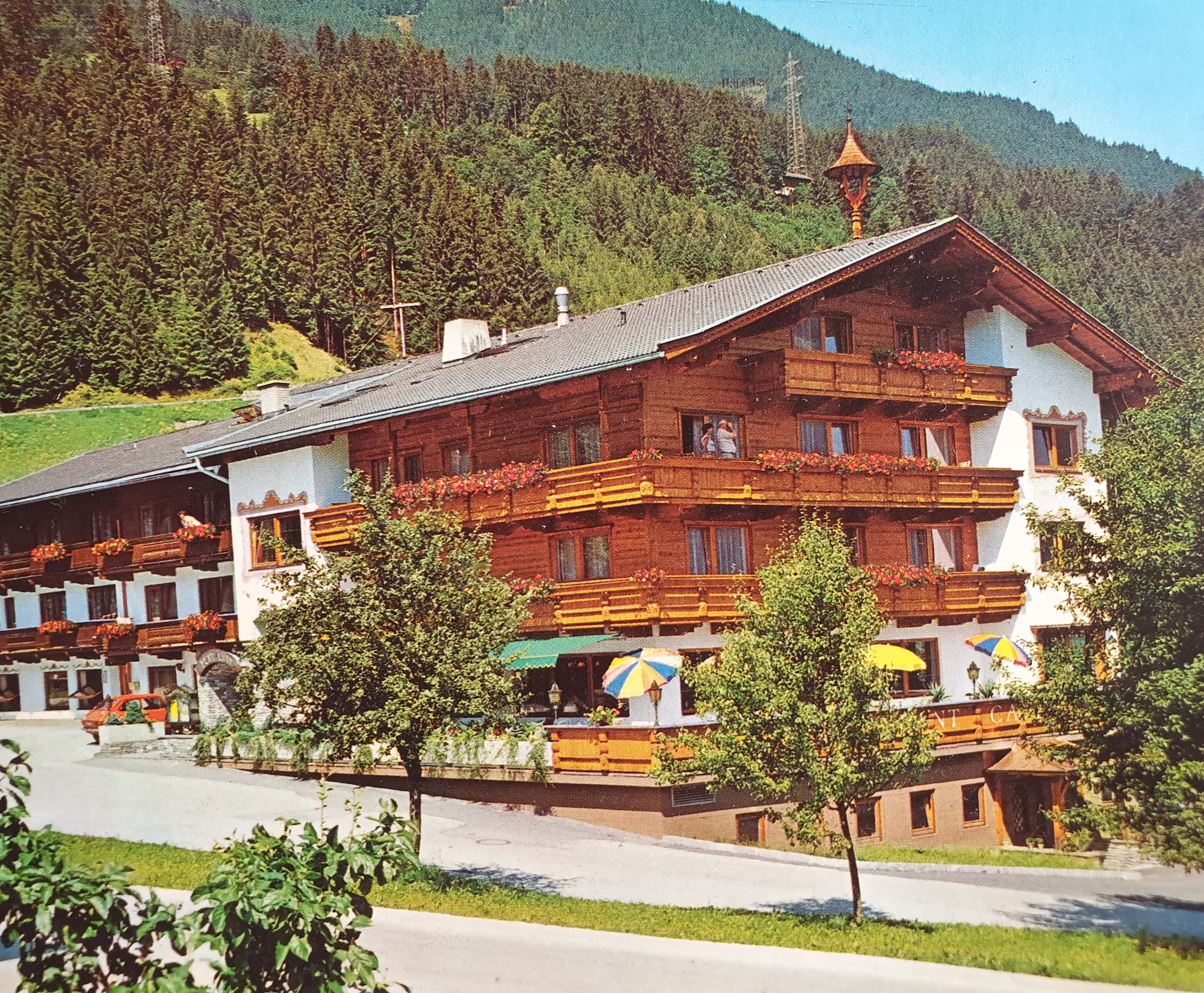 Der Platzlhof im Jahr 1978 Ein traditionelles alpines Hotel mit Holzverkleidung, Balkonen und bunten Sonnenschirmen, umgeben von Bäumen und Bergen im Hintergrund.