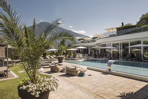 Sonniger Poolgarten des Hotel Platzlhof mit Außenpool, Liegeflächen, Palmen und Blick auf die Zillertaler Berge.
