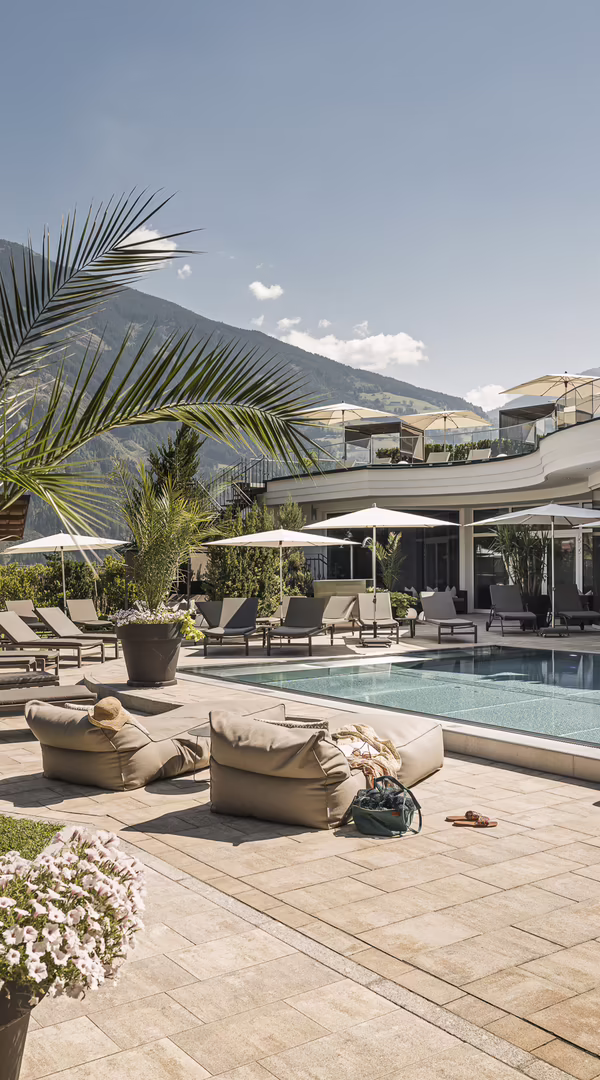 Sonniger Poolgarten des Hotel Platzlhof mit Außenpool, Liegeflächen, Palmen und Blick auf die Zillertaler Berge.