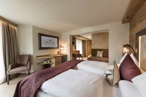Modernes Hotelzimmer mit zwei Einzelbetten, einem Stuhl, einem Tisch und einem Fenster mit Vorhängen. Angenehme Farbgestaltung und warmes Licht.