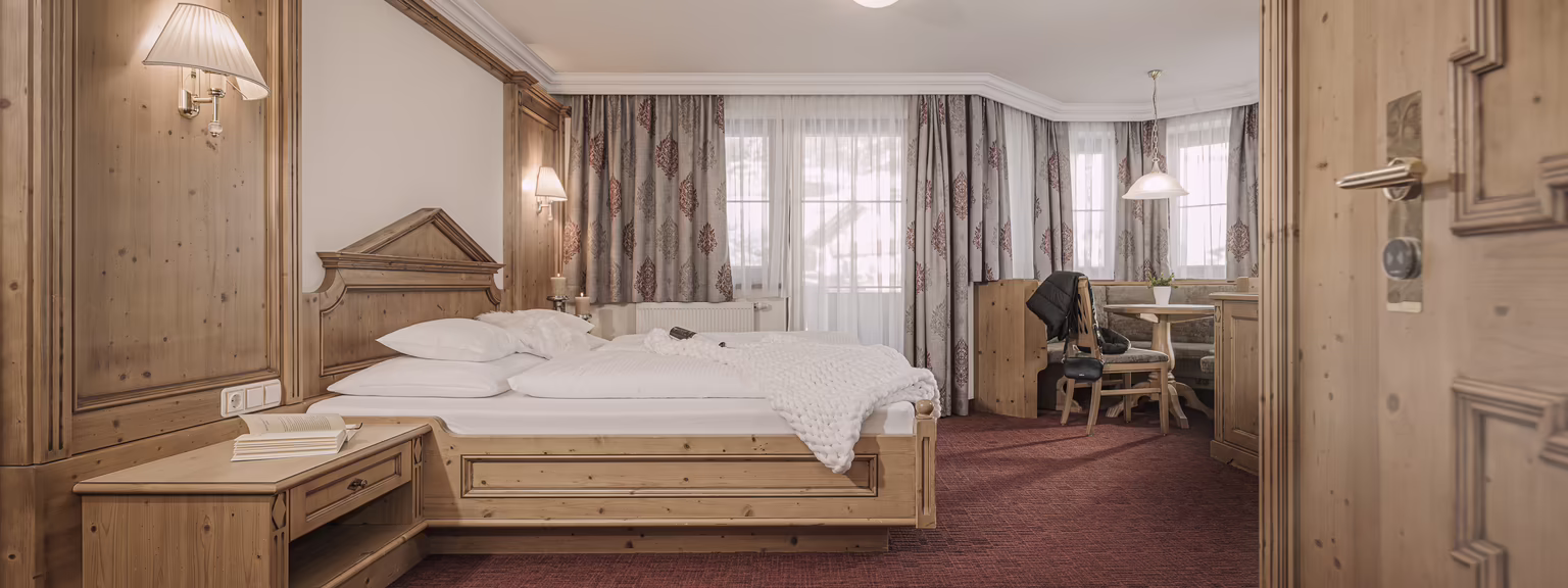 Gemütliches Hotelzimmer mit Holzdecken, Doppelbett, Schreibtisch und Fenster mit Vorhängen. Rote Teppichboden und warmes Licht.