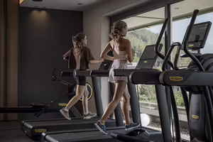 Zwei junge Frauen joggen auf Laufbändern in einem modernen Fitnessraum mit Fenstern und Blick ins Freie.