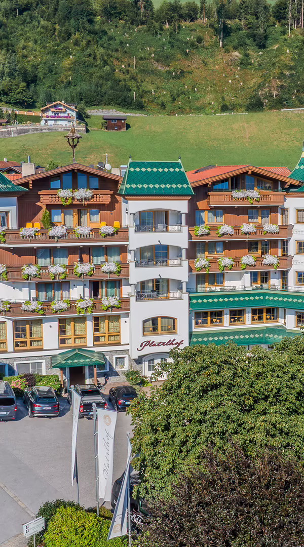 Hotel Platzlhof mit grünem Dach und Balkonen, umgeben von einer grünen Landschaft und Bäumen. Im Vordergrund sind Parkplätze mit mehreren Autos.
