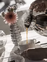 Eine Person gießt dampfenden Kaffee aus einer Kaffeekanne in eine Tasse, während eine blühende Vase im Hintergrund steht.