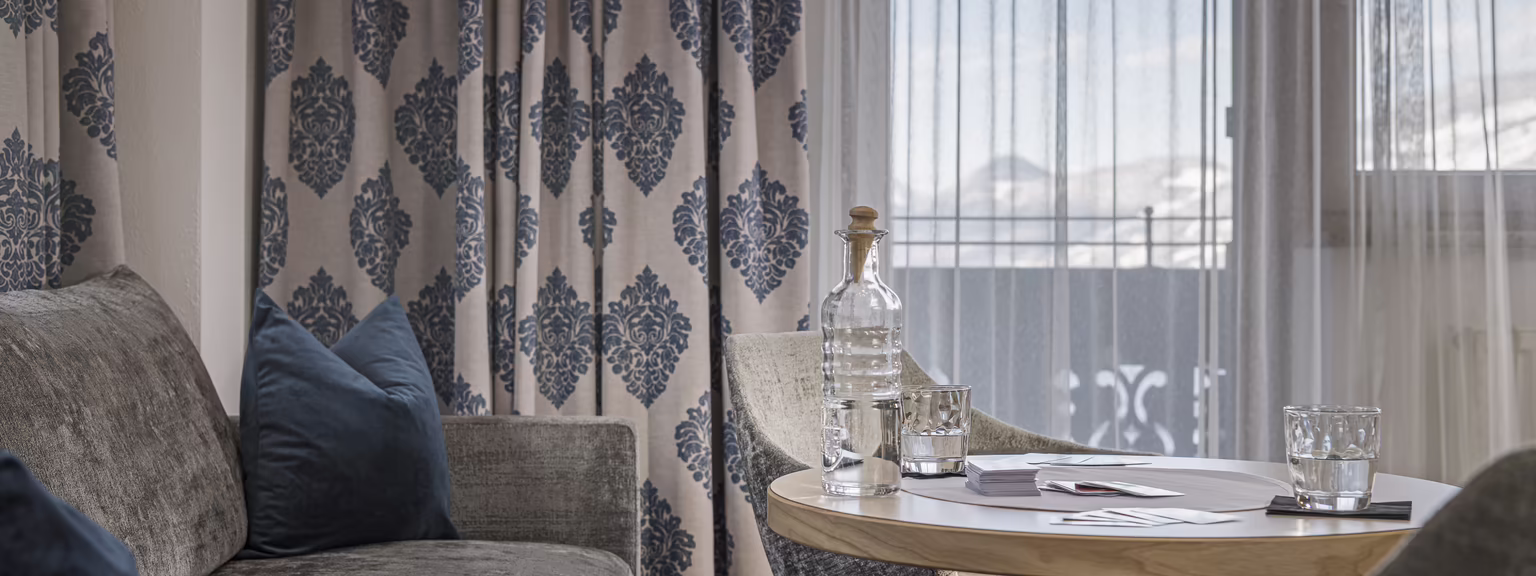 Alpiner Wohnkomfort in der Wohlfühlsuite Rosalie Helle Sitzecke im Hotel Platzlhof mit Sofa, Tisch und Blick durch die Balkontür auf die Zillertaler Bergwelt.