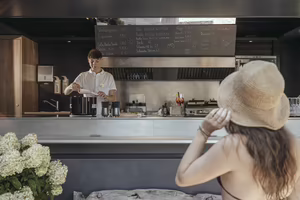 Ein Bartender in einem Café bereitet Getränke vor, während eine Frau im Hintergrund mit einem Strohhut zuschaut. Blumen stehen auf dem Tisch.