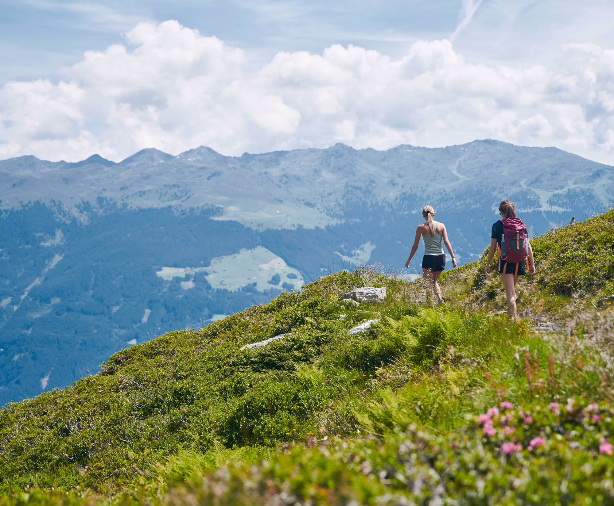 Zwei Personen wandern auf einem Pfad in bergiger Landschaft. Im Hintergrund sind hohe Berge und grüne Wiesen zu sehen. Der Himmel ist blau mit einigen Wolken.