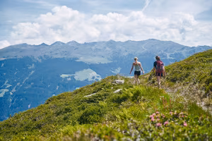 Zwei Personen wandern auf einem Pfad in bergiger Landschaft. Im Hintergrund sind hohe Berge und grüne Wiesen zu sehen. Der Himmel ist blau mit einigen Wolken.