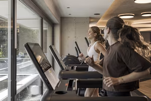 Zwei Frauen laufen auf Laufbändern in einem modernen Fitnessstudio mit großen Fenstern und natürlichem Licht.