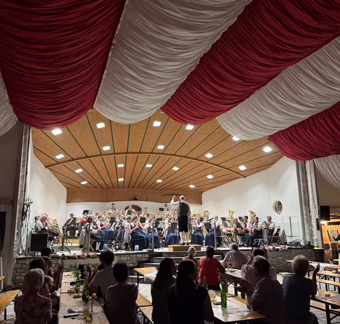 Platzkonzert der Bundesmusikkapelle Ried Kaltenbach Musiker der Bundesmusikkapelle Ried Kaltenbach spielen auf einer Bühne, während Publikum an Biertischen sitzt, umgeben von roter und weißer Dekoration.