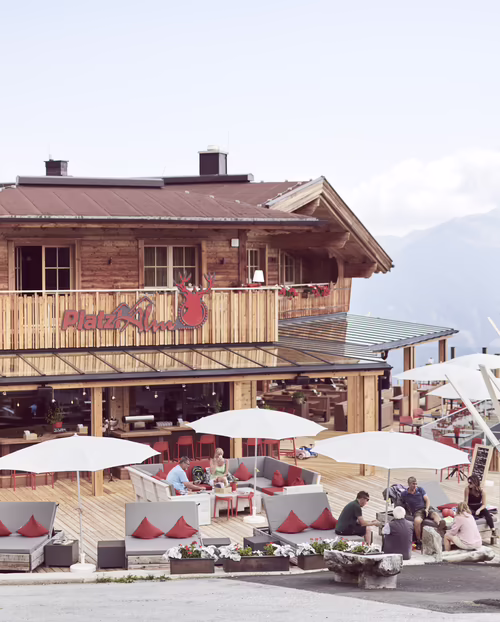 Platzlalm im Sommer Ein Holzhaus mit großer Terrasse, auf der Gäste unter Sonnenschirmen sitzen. Im Hintergrund sind Berge und ein blauer Himmel zu sehen.