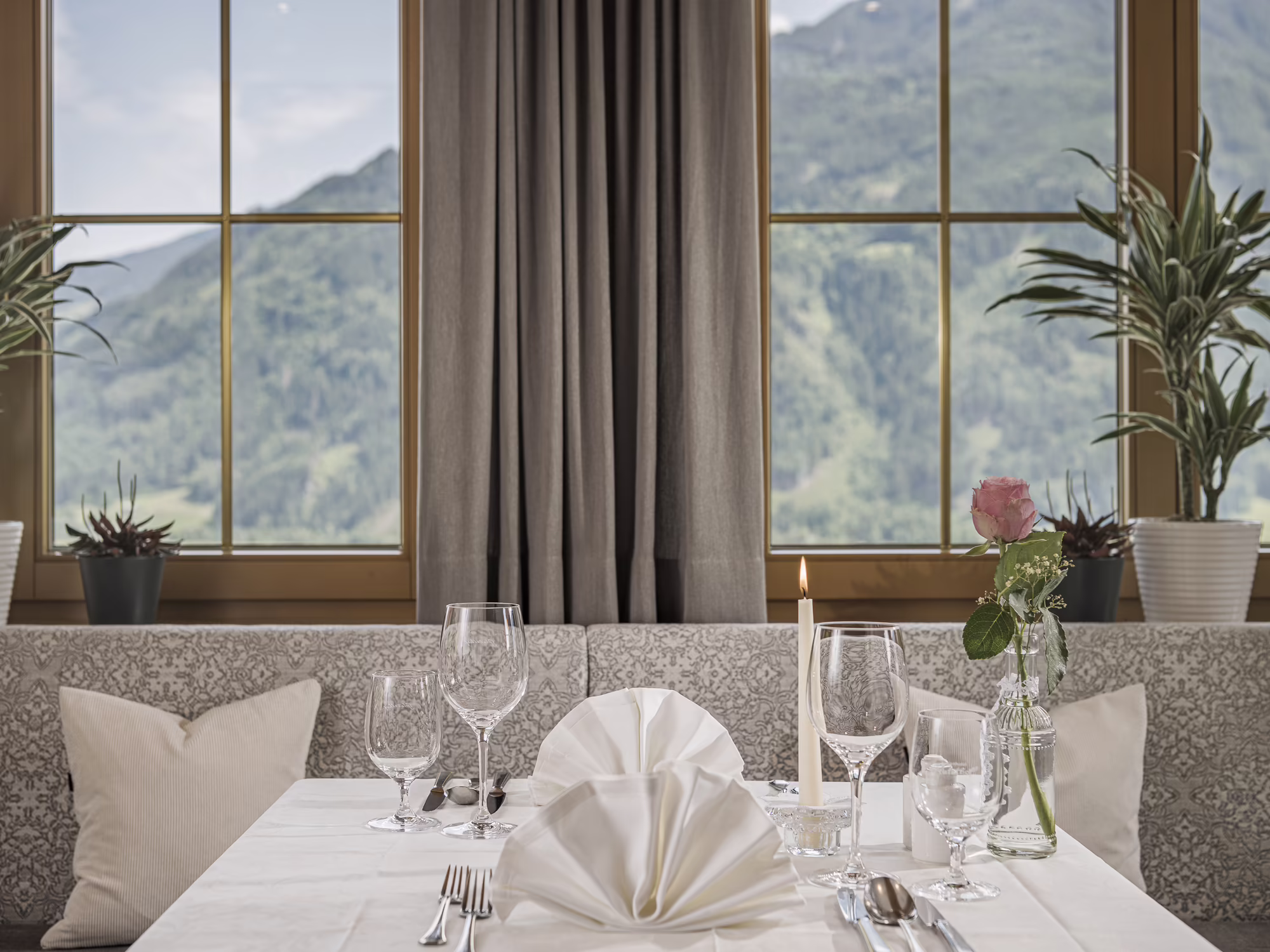 Ein eleganter Tisch mit weißer Tischdecke, Gläsern und einer Rose, mit Blick auf grüne Berge durch große Fenster.