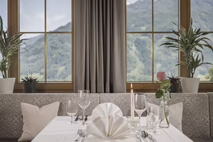 Ein eleganter Tisch mit weißer Tischdecke, Gläsern und einer Rose, mit Blick auf grüne Berge durch große Fenster.