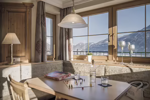 Gemütlicher Essbereich mit Tisch, Spiel und Getränken. Große Fenster mit Blick auf eine winterliche Berglandschaft.