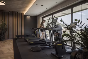 Fitnessraum mit modernen Geräten und einer Person, die auf einem Laufband trainiert, vor großen Fenstern mit Blick ins Freie.