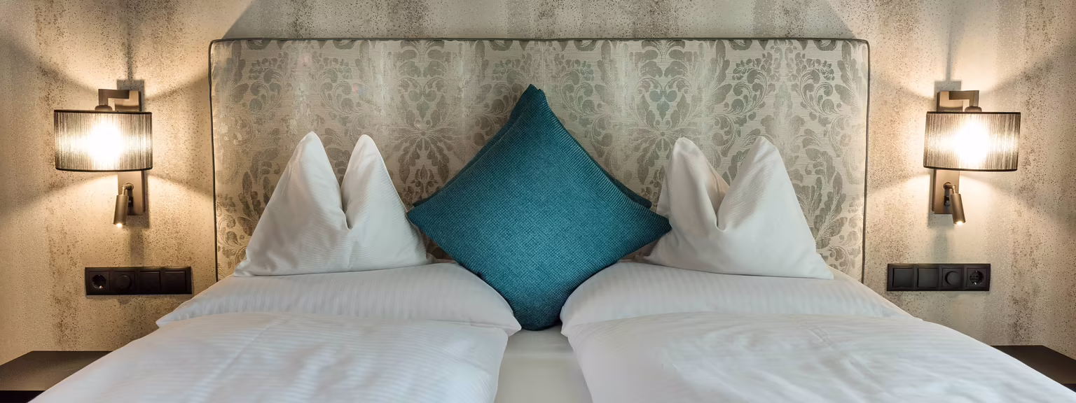 Doppelbett mit dekorativen Kopfteil, Kissen und einem blauen Zierkissen; beleuchtet von zwei Wandlampen.