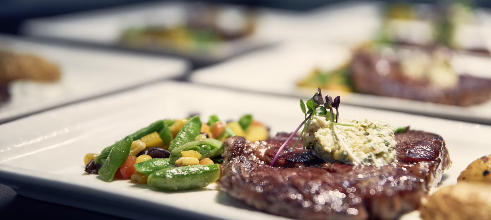 Ein Teller mit saftigem Steak, garniert mit Kräuterbutter, und einer Beilage aus buntem Gemüse. Weitere Teller im Hintergrund.