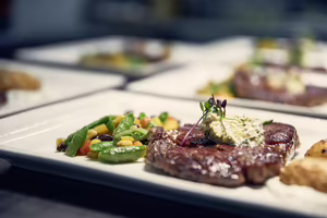 Ein Teller mit saftigem Steak, garniert mit Kräuterbutter, und einer Beilage aus buntem Gemüse. Weitere Teller im Hintergrund.