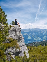 Person sitzt auf einer Felsenkante und blickt auf eine Berglandschaft mit Hügeln und Wolken am Himmel.