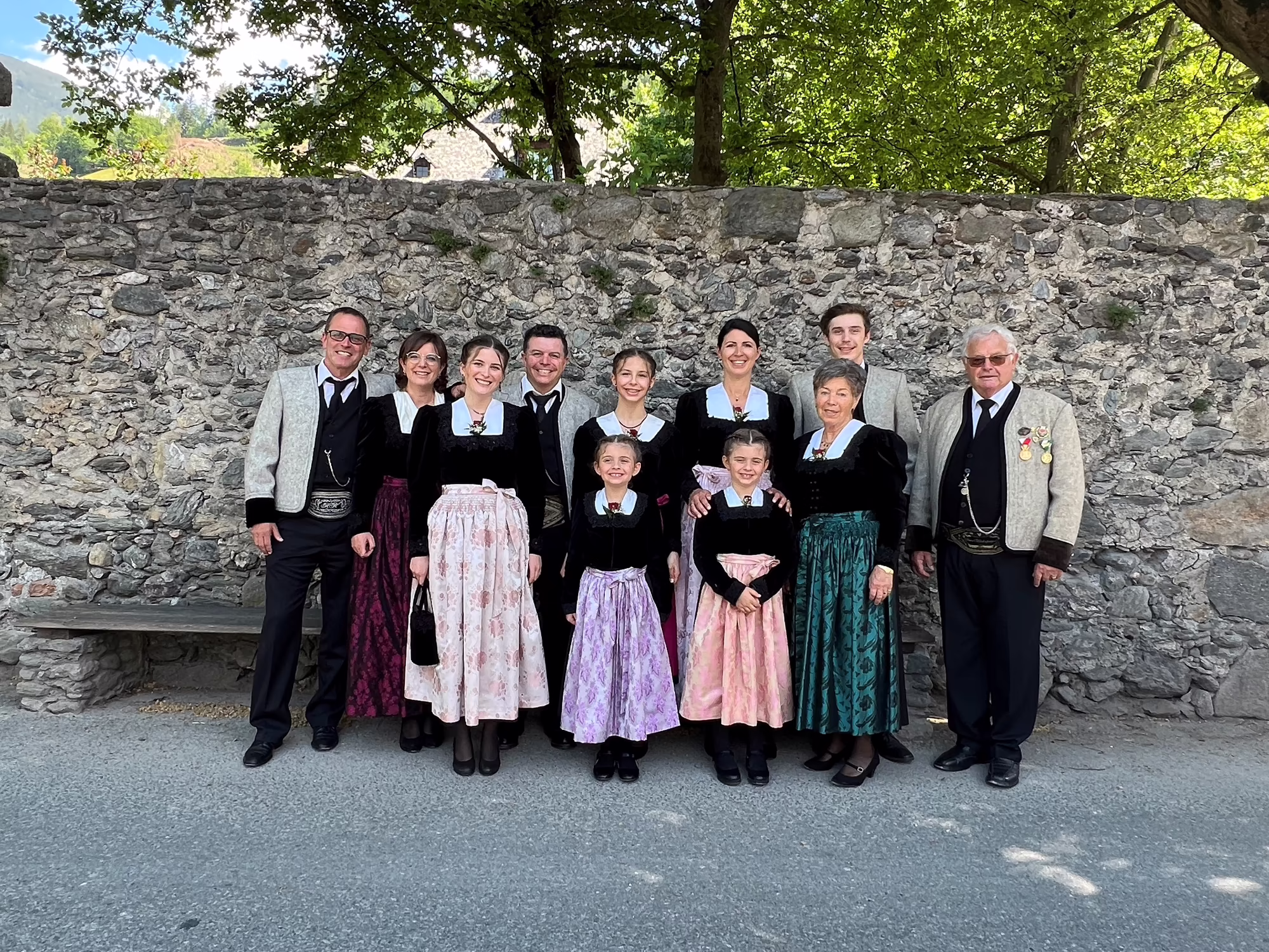 Familie Wimpissinger - Hotel Platzlhof in Ried im Zillertal Familie Wimpissinger in traditioneller Tracht steht vor einer Steinmauer und lächelt in die Kamera. Bäume sind im Hintergrund sichtbar.