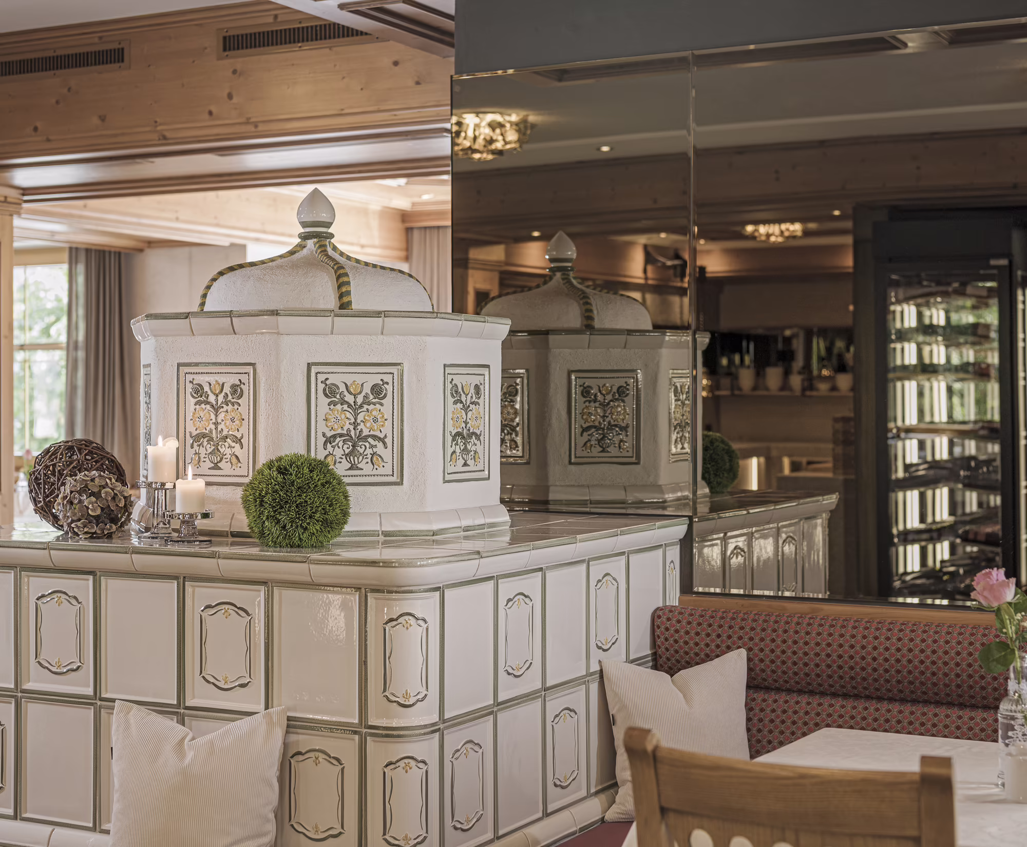 Kachelofen im Restaurant im Hotel Platzlhof Moderne Gaststube mit einem weißen Kachelofen, gepolsterter Bank und einem großen Spiegel an der Wand.